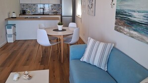 A modern living room with a blue sofa, a dining table with chairs, a kitchen area with white cabinets, and a large painting of a seascape.