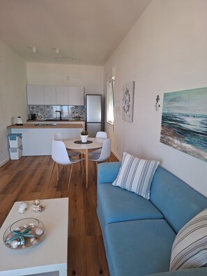 Un salon moderne avec un canapé bleu, une table à manger avec des chaises, un coin cuisine doté de meubles blancs et un grand tableau représentant un paysage marin.