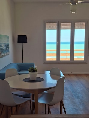 Une pièce moderne avec une table de salle à manger ronde, des chaises blanches, un canapé bleu, une lampe sur pied et une grande fenêtre donnant sur la plage.