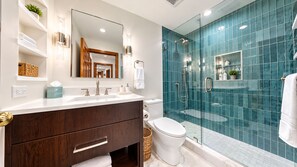 Un baño moderno con ducha de vidrio, lavamanos blanco con tocador de madera y un espejo grande sobre el tocador.