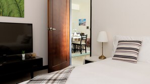 Quarto de hotel com cama, televisão, secretária e zona de refeições visível por uma porta aberta.