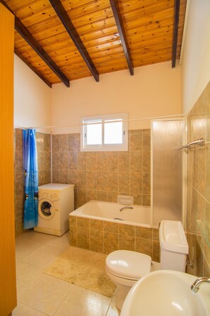 A bathroom with a wooden ceiling, a washing machine, a bathtub, a toilet, and a window. - Corallia Villa ,3 Bedrooms see and Forest View (Ayia Marina Chrysochous)