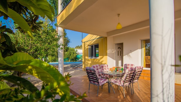 A patio with a table and chairs, a yellow building in the background, and greenery in the foreground. - Corallia Villa ,3 Bedrooms see and Forest View (Ayia Marina Chrysochous)