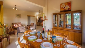 A dining area with a wooden table set for four, surrounded by chairs. There is a cabinet with glass doors to the right, and a painting above it. - Corallia Villa ,3 Bedrooms see and Forest View (Ayia Marina Chrysochous)