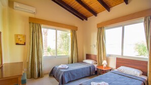A hotel room with two beds, a wooden desk, and a chair. There are curtains on the windows, and an air conditioning unit is mounted on the wall. - Corallia Villa ,3 Bedrooms see and Forest View (Ayia Marina Chrysochous)