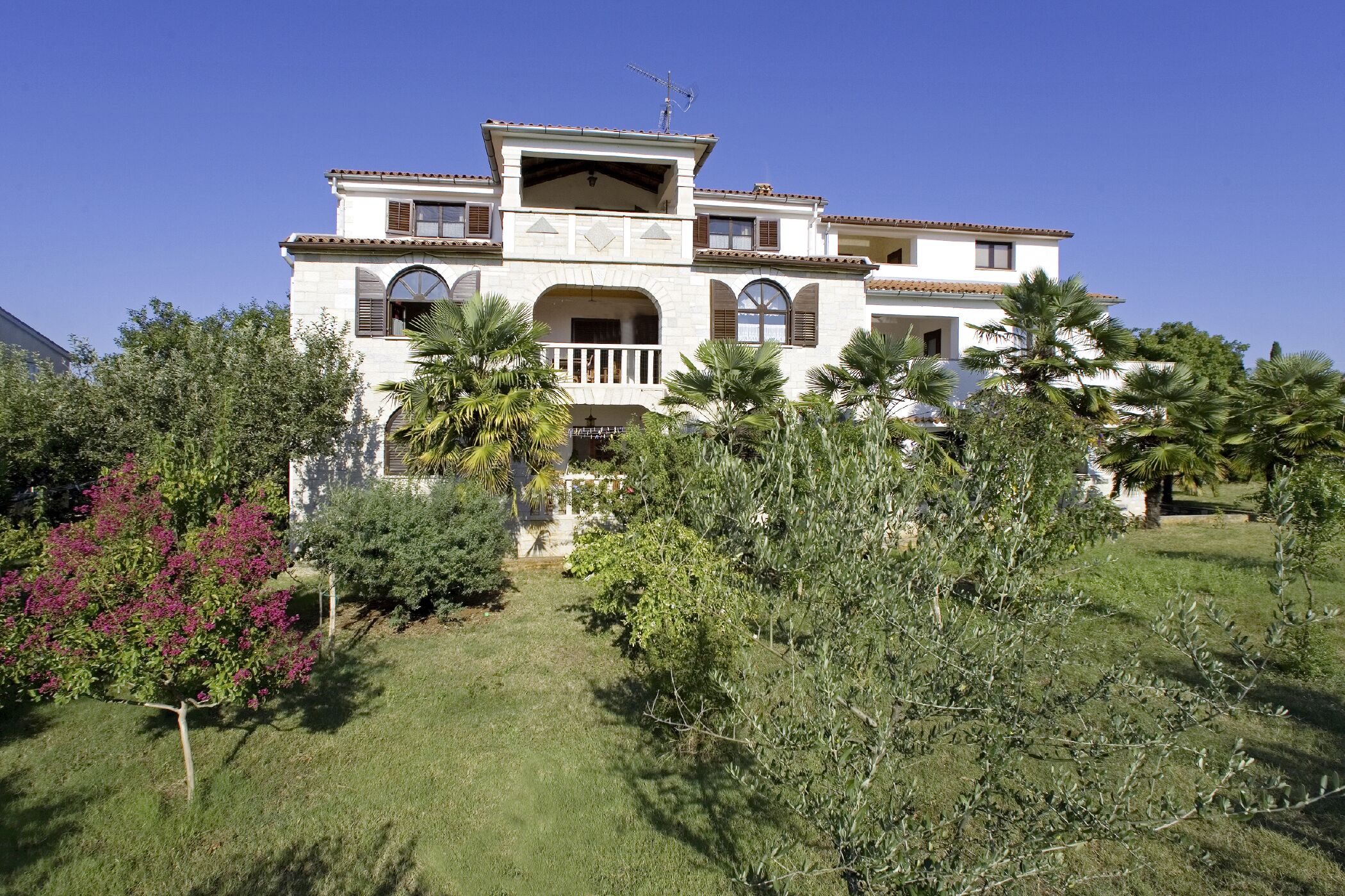 A two-story house with a balcony, surrounded by lush greenery and palm trees.