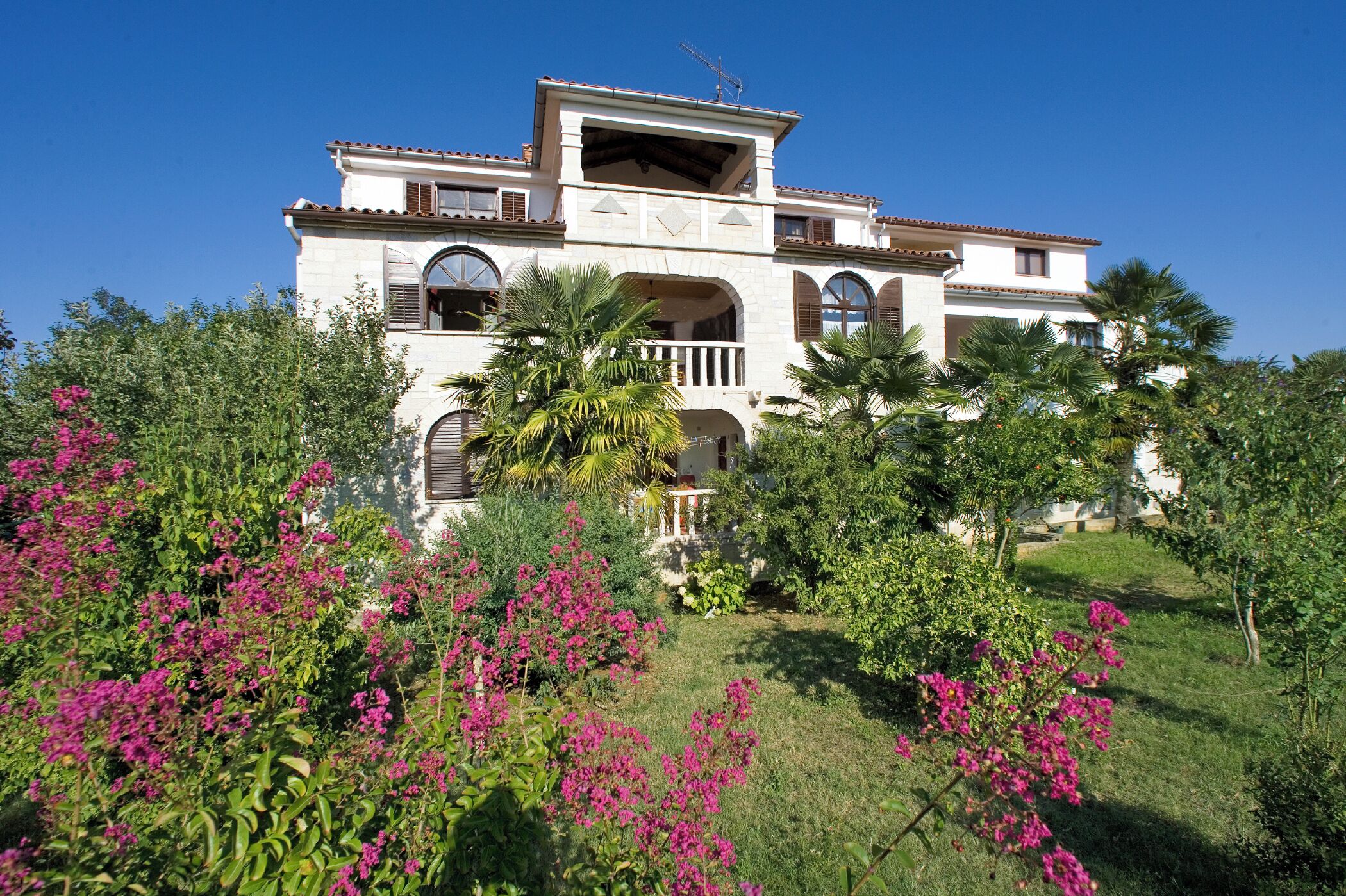 Un edificio grande de dos pisos con varios balcones y ventanas arqueadas, rodeado de abundante follaje y plantas con flores.