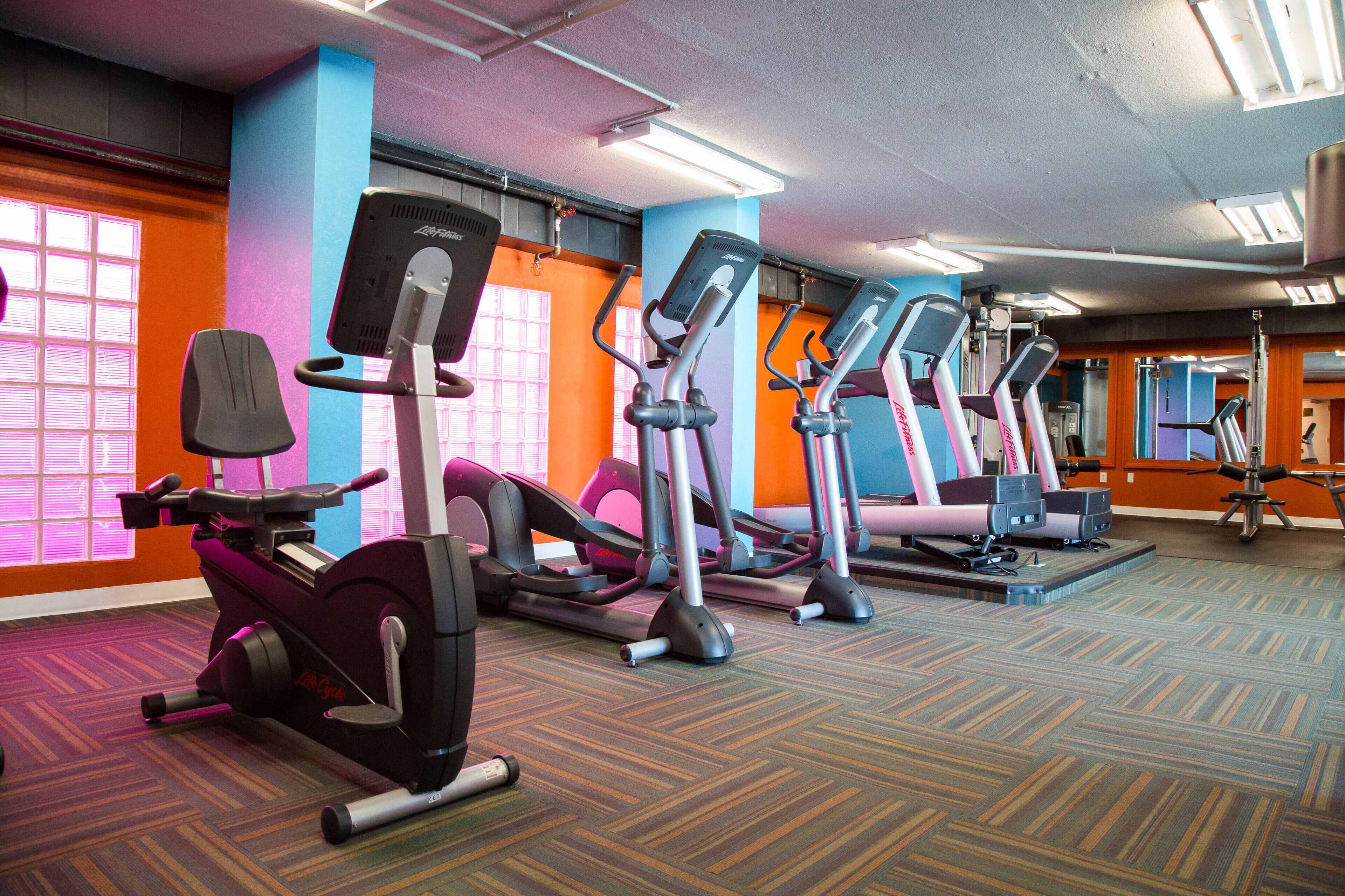 A gym with stationary bikes and elliptical machines, carpeted flooring, and colorful walls.