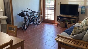 Een woonkamer met een houten bank, een televisie op een houten dressoir, een fiets en een raam met uitzicht op groen.