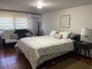 Une chambre à coucher avec un lit, une chaise, une table de chevet, une lampe et une fenêtre avec des stores.