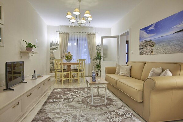 A living room with a beige sofa, a TV on a cabinet, a dining table with chairs, and a chandelier.
