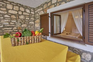 A room with a stone wall, a wooden-framed window with curtains, a wooden crate with vegetables, and a yellow bedspread.