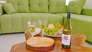 A green sofa with cushions, a wooden table with a wine bottle and glasses, and a plate with grapes and cheese.