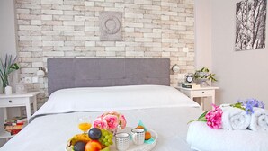 A neatly made bed with a grey headboard, a tray of fruit and flowers, and a rolled towel on the side.
