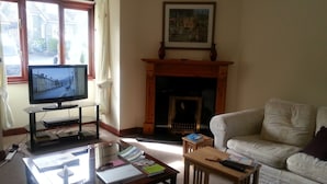 A living room with a television on a stand, a fireplace, a sofa, and a coffee table with books and a remote control. - Spacious  holiday cottage in Bowness on Windermere, Lake Distrct, Uk (Windermere)