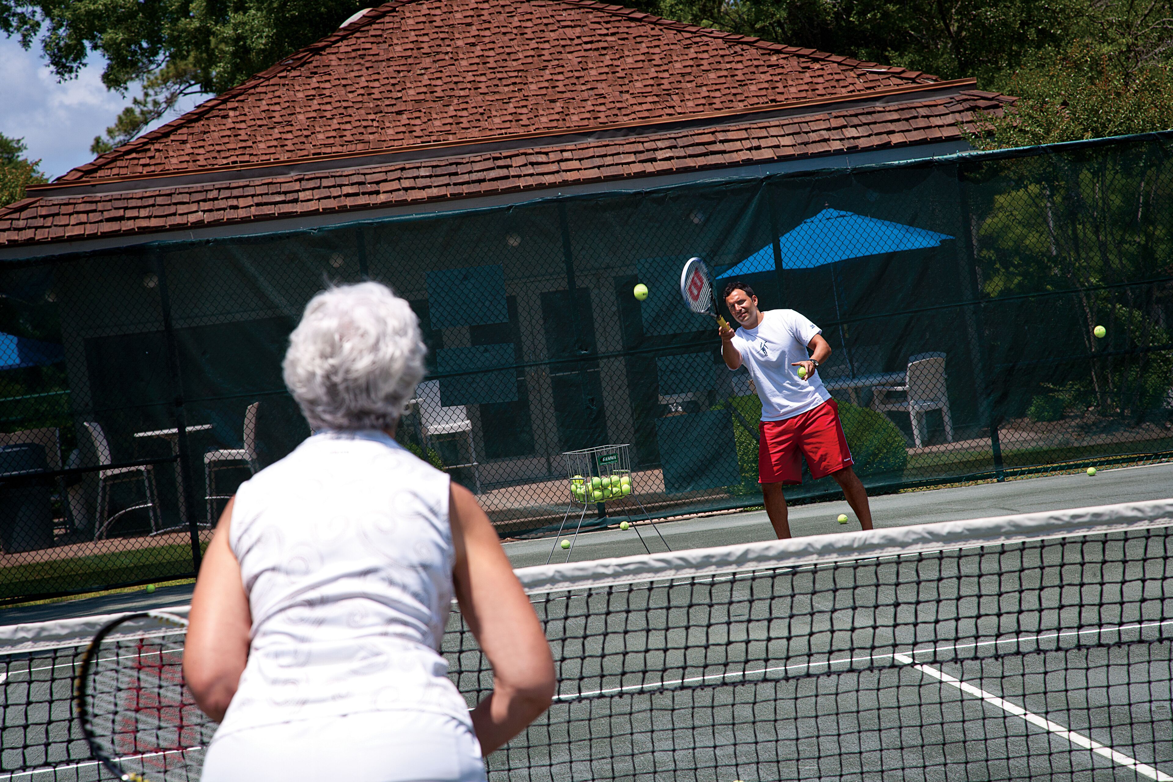 A tennis court with a net, a building with a red roof, and a fence.