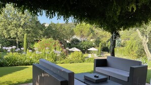 Zona salotto esterna con divano e sedie, circondata da un giardino ricco di piante e fiori.