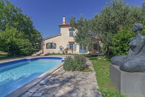 A pool with a stone deck, a statue, and a house with a tiled roof and shutters.