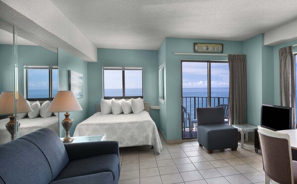 A hotel room with a bed, a sofa, a chair, a TV, and a balcony with a view of the sea.
