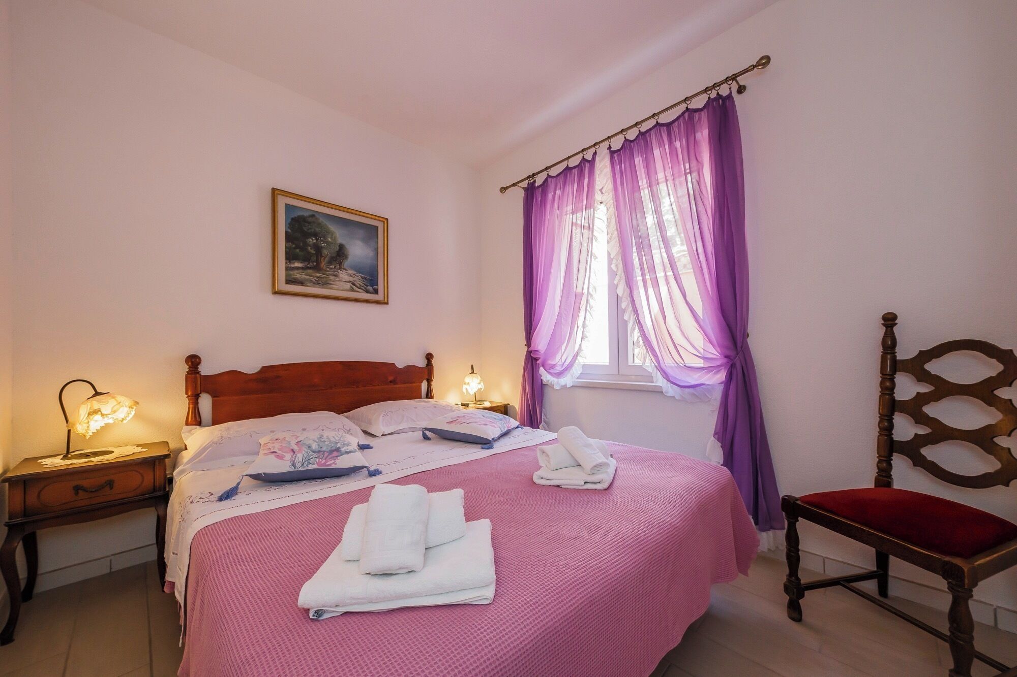 A bedroom with a wooden bed, a nightstand with a lamp, a chair, and a window with purple curtains.
