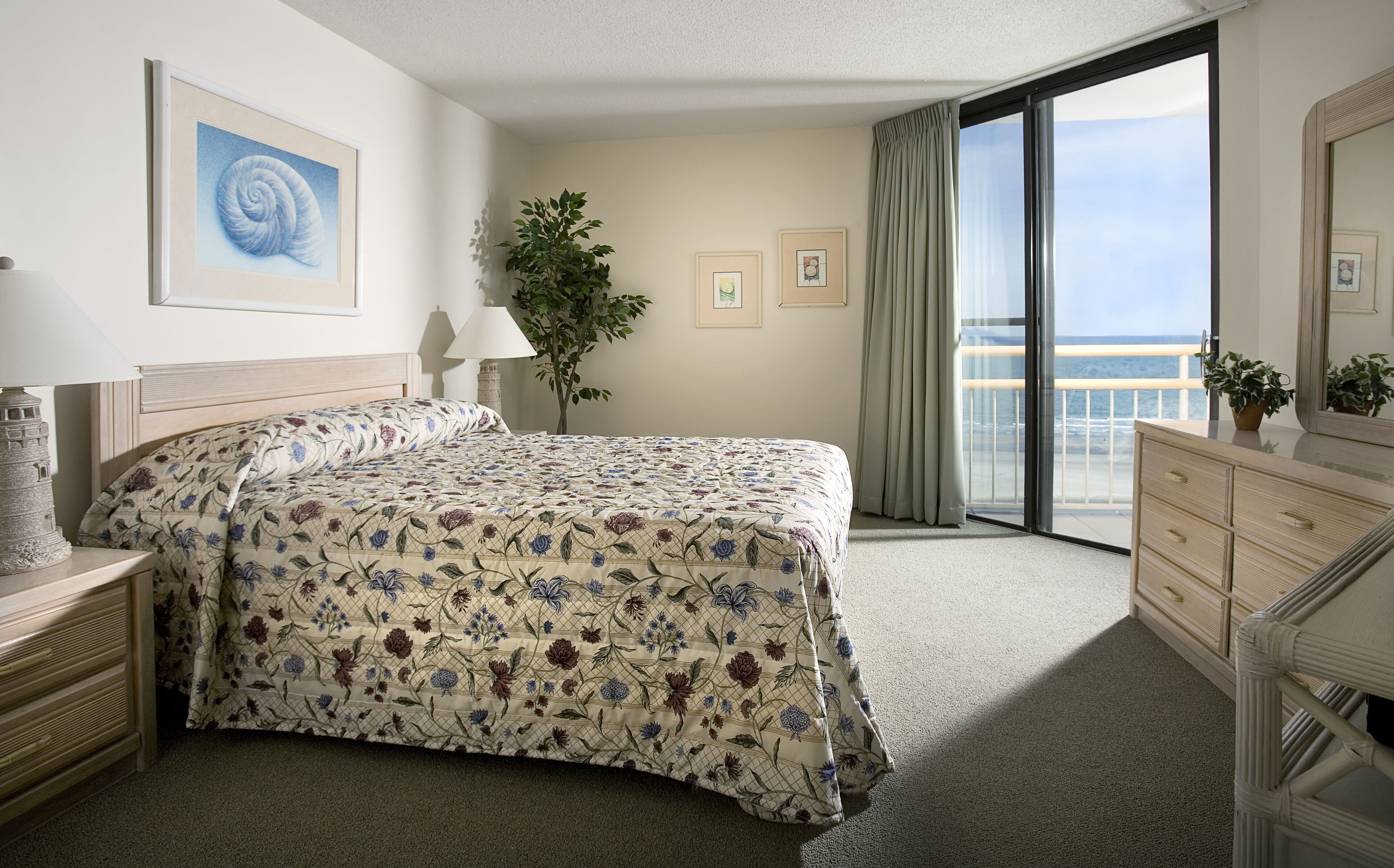 A hotel room with a bed, a dresser, a nightstand, a lamp, a plant, and a large window with a view of the ocean.