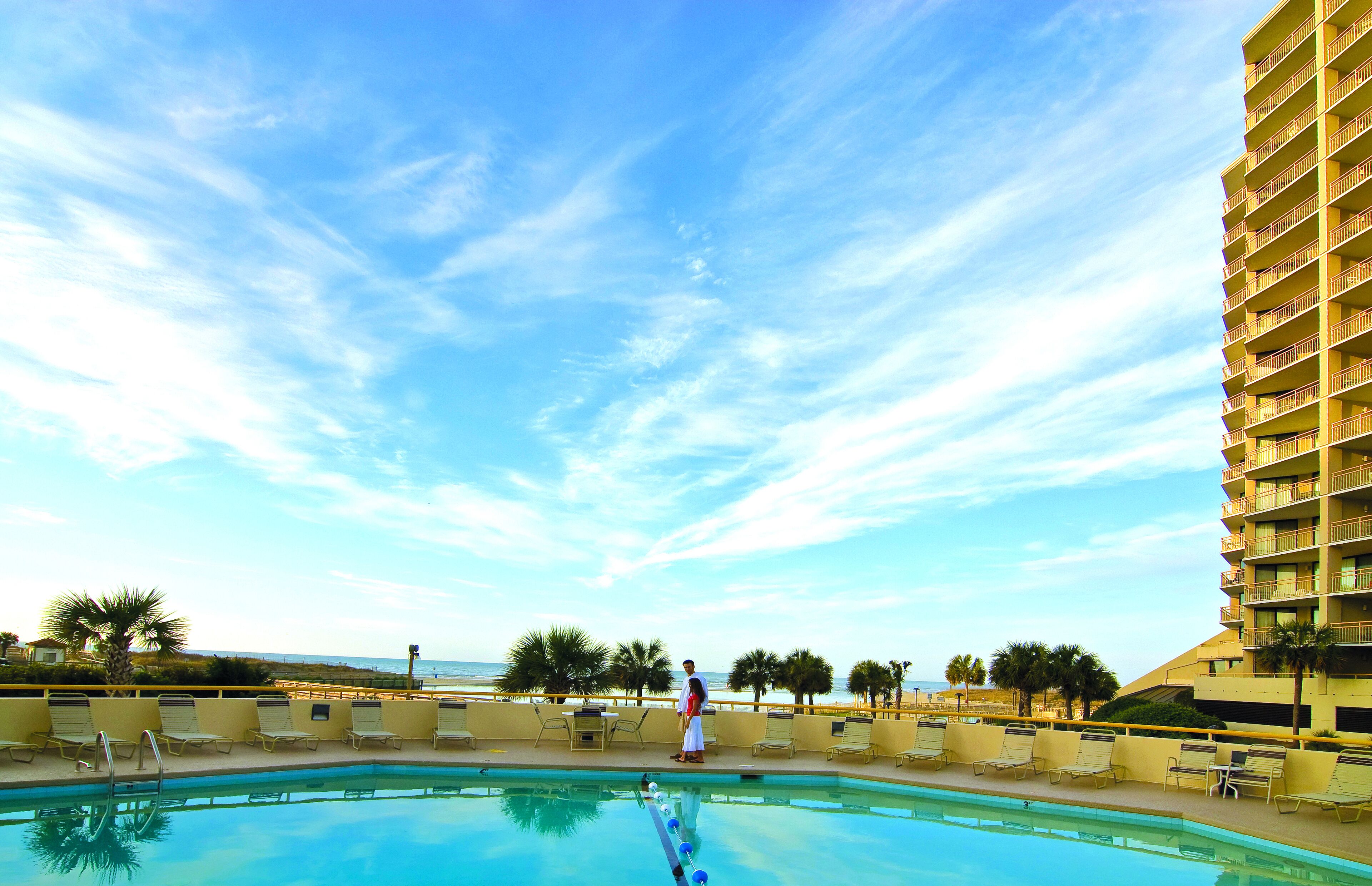 Zona de piscina bajo un cielo despejado, una persona de pie junto a la piscina y un rascacielos al fondo.