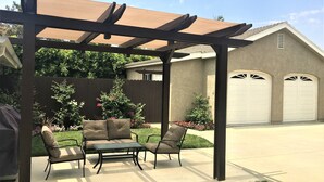 Ein überdachter Patio-Bereich mit zwei gepolsterten Stühlen und einem kleinen Tisch, eine Pergola darüber, und ein Haus mit Garage im Hintergrund.