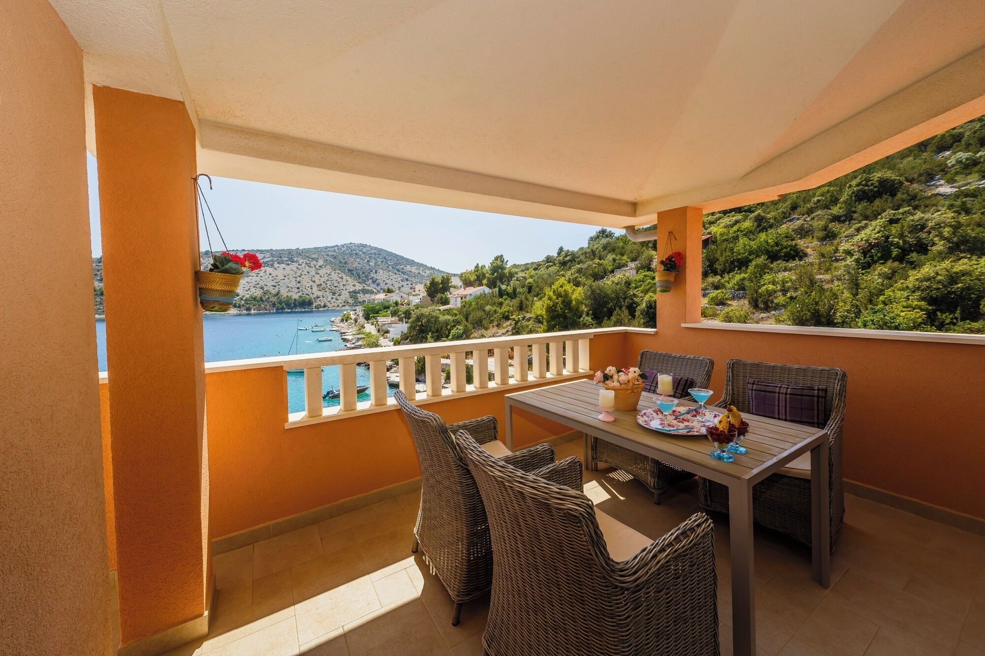 A balcony with wicker furniture, a table set with a drink, and a view of a lake and hills.