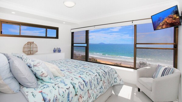 A bedroom with a large window overlooking a beach, a bed with a blue and white fish-patterned bedspread, a white sofa, and a flat-screen TV mounted on the wall.