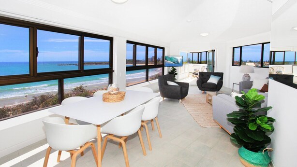 Una habitación moderna con un amplio ventanal con vista a la playa, una zona de comedor con sillas blancas y un espacio de estar con un sofá y sillones grises.