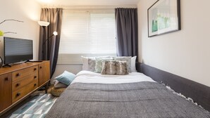 Ein Schlafzimmer mit Bett, Schminktisch, Lampe, einem Fenster mit Vorhängen und einem gerahmten Bild an der Wand.