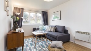 Ein modernes Wohnzimmer mit einem grauen Sofa, einem Holztisch, einem gemusterten Teppich und einem großen Fenster mit Vorhängen.