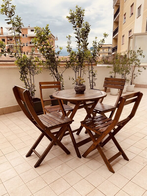 Una mesa redonda de madera con cuatro sillas en una terraza con baldosas, rodeada de macetas con plantas.