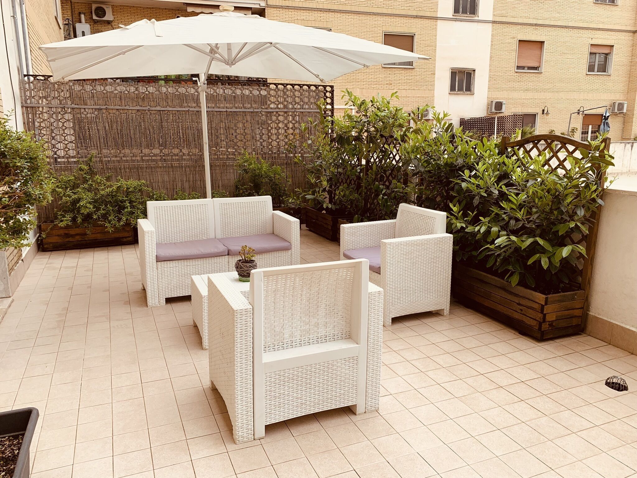 En patio med hvide fletmøbler, herunder en sofa og stole, under en stor hvid parasol. Der er plantede krukker og et træhegn i baggrunden.