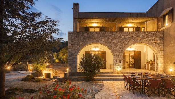 A stone building with two arched entrances, a patio with a table and chairs, and a garden with red flowers. - Luxurious Mani Peninsula Traditional Tower (East Mani)