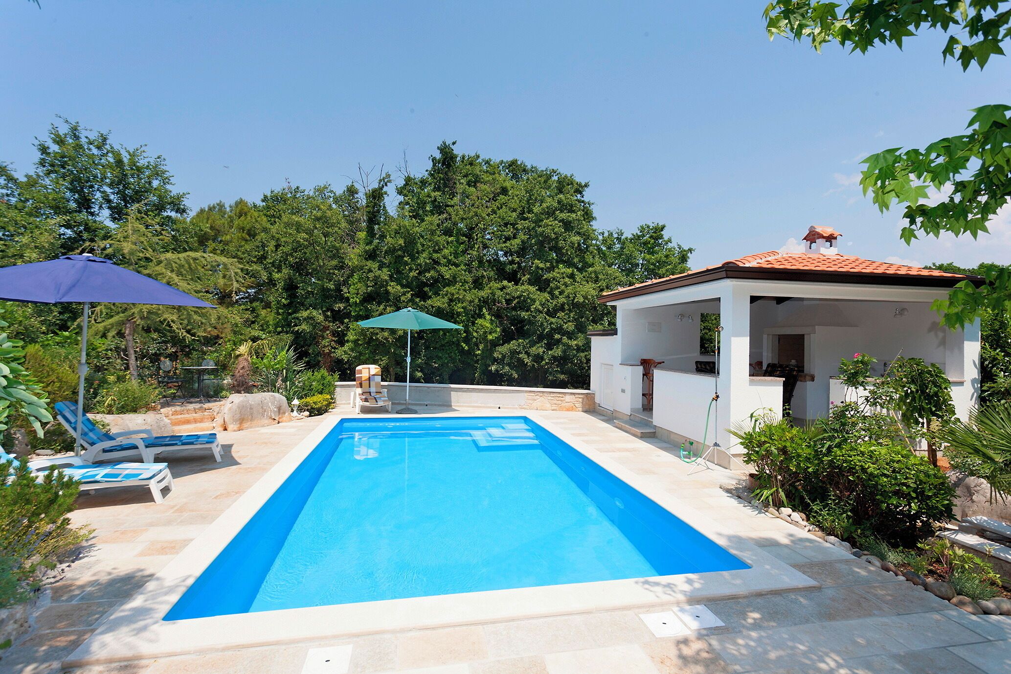 Piscina retangular com duas espreguiçadeiras e guarda-sóis, rodeada por vegetação e um pequeno pavilhão.