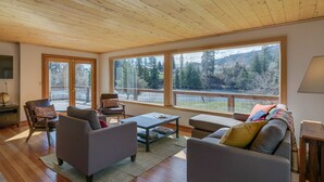 A spacious living room with a high ceiling, large windows offering a view of a forested area, and a cozy seating arrangement. - Peshastin River Place-Riverfront near Leavenworth (Peshastin)