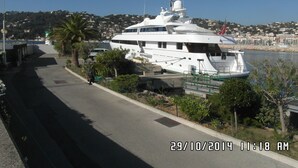 Un yate blanco grande atracado en un puerto deportivo, con vistas claras a los alrededores, incluyendo una carretera y edificios al fondo.
