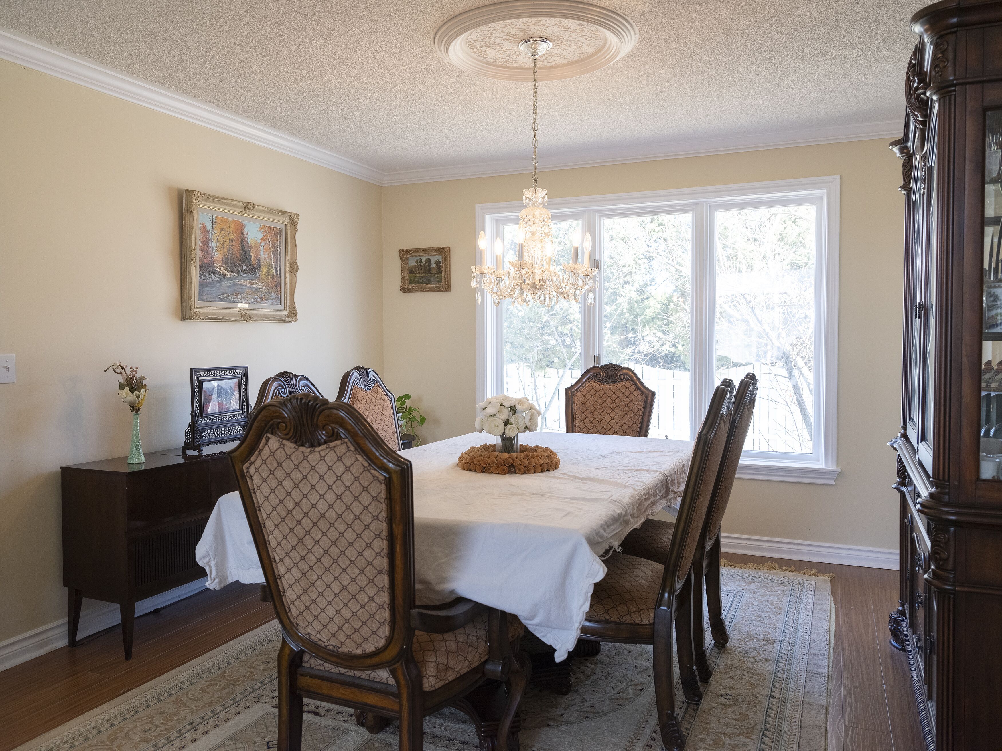 A dining room with a table set for six, surrounded by wooden chairs with cushioned seats. A chandelier hangs from the ceiling, and there are framed pictures on the walls.