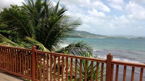 Eine Holzterrasse mit Metallgeländer, die auf einen Strand mit Palmen und eine Aussicht auf ferne Hügel blickt.