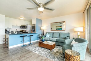 A modern living room with a blue sofa, a wooden coffee table, and a kitchen area with white cabinets and a bar stool.