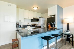 Una cucina moderna con mobili bianchi, un piano di lavoro nero e una parete blu decorativa. Sono presenti due sgabelli da bar e un piccolo tavolo con una ciotola di frutta.
