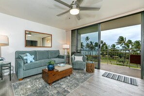 Un soggiorno moderno con un divano grigio, un tavolino in legno e un tavolo in vetro con una lampada. Un grande specchio appeso al muro, e un balcone con vista su palme.