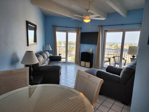 A living room with a sofa set, a TV, a ceiling fan, and a balcony with a view. - Sunsets & Savings​​​​​​​ • Pool & Hot Tub • Beachfront with Bay Views (South Padre Island)