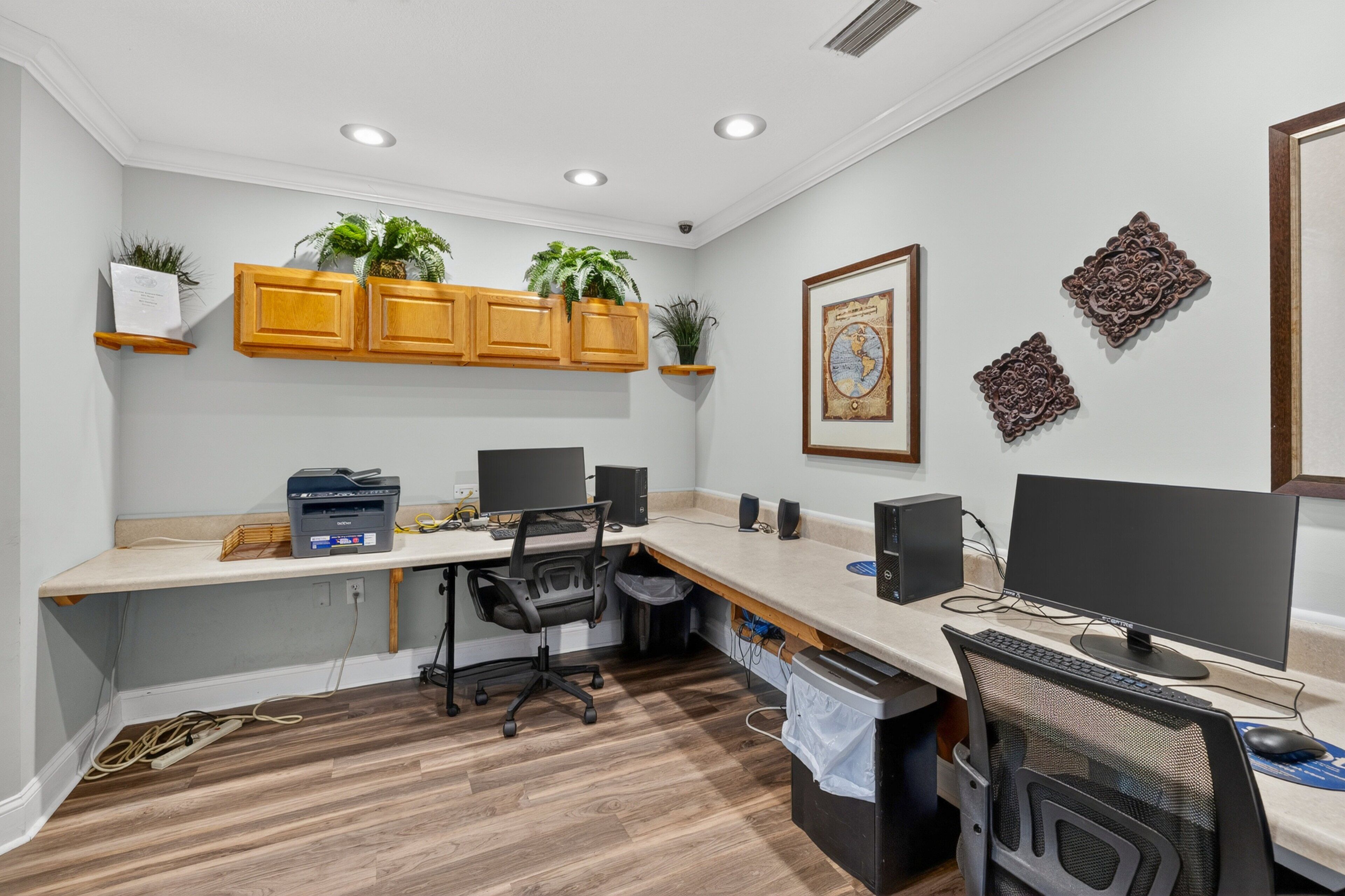A modern office with a long desk, two monitors, a computer, and a chair. There are wooden cabinets above the desk and potted plants on the shelves.