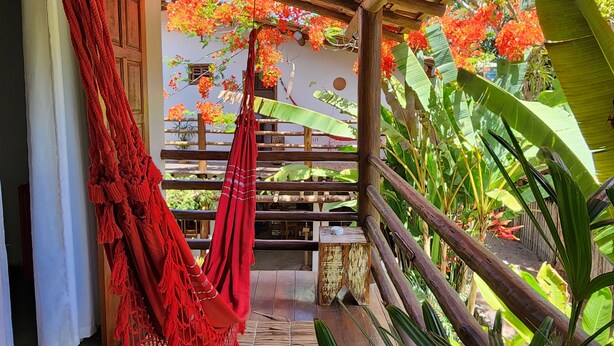 A wooden balcony with a red hammock, a wooden railing, and lush green foliage. - Hostel e Pousada Casa de Paixão (Porto Seguro)