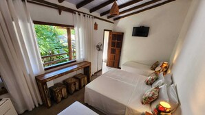 A room with two beds, a bench, a TV, and a window with a view of greenery. - Hostel e Pousada Casa de Paixão (Porto Seguro)