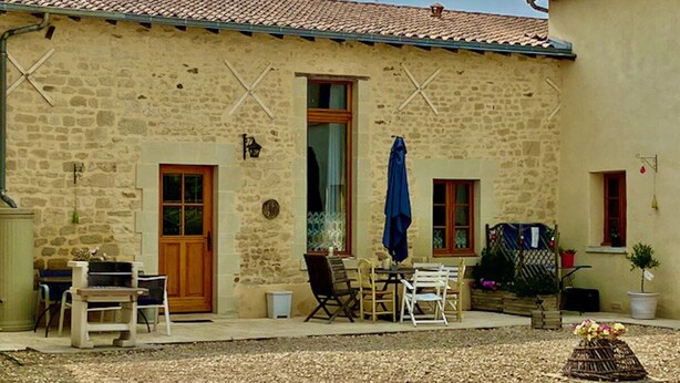Un edificio de piedra con techo de tejas, una puerta de madera y una ventana con cortina. Hay una mesa con sillas y una sombrilla azul.