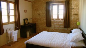 Un dormitorio con pared de piedra, una cama con ropa de cama blanca, un escritorio con silla y una ventana con cortinas.