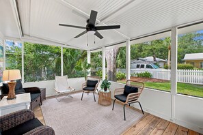 A covered outdoor area with wicker furniture, a ceiling fan, and large windows offering a view of the surrounding greenery and houses.